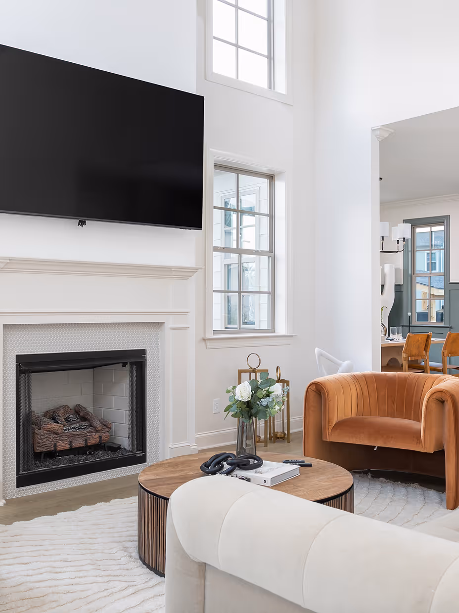 Bright living room with a mounted flat-screen TV above a white fireplace, a round wooden coffee table with a vase of flowers, and a brown velvet armchair.