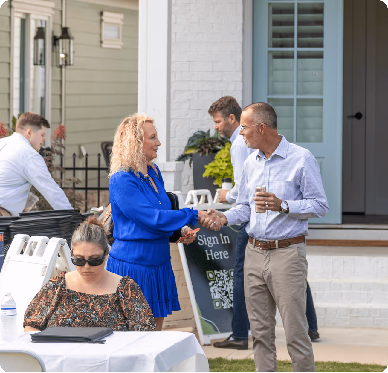 Two people shaking hands outdoors near a sign that says 'Sign In Here' with a QR code, while others are engaged in various activities nearby.