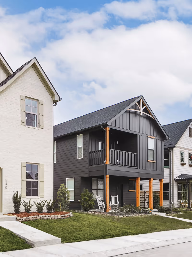 Modern two-story house with dark exterior siding, wooden columns, a balcony, rocking chairs on a porch, and a lawn with landscaping.