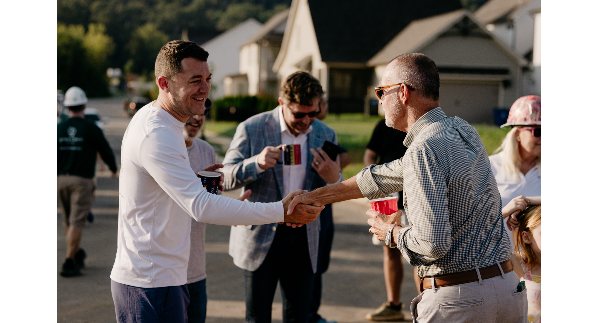 Two men smiling and shaking hands outdoors, each holding a drink, with a group of people and houses in the background.