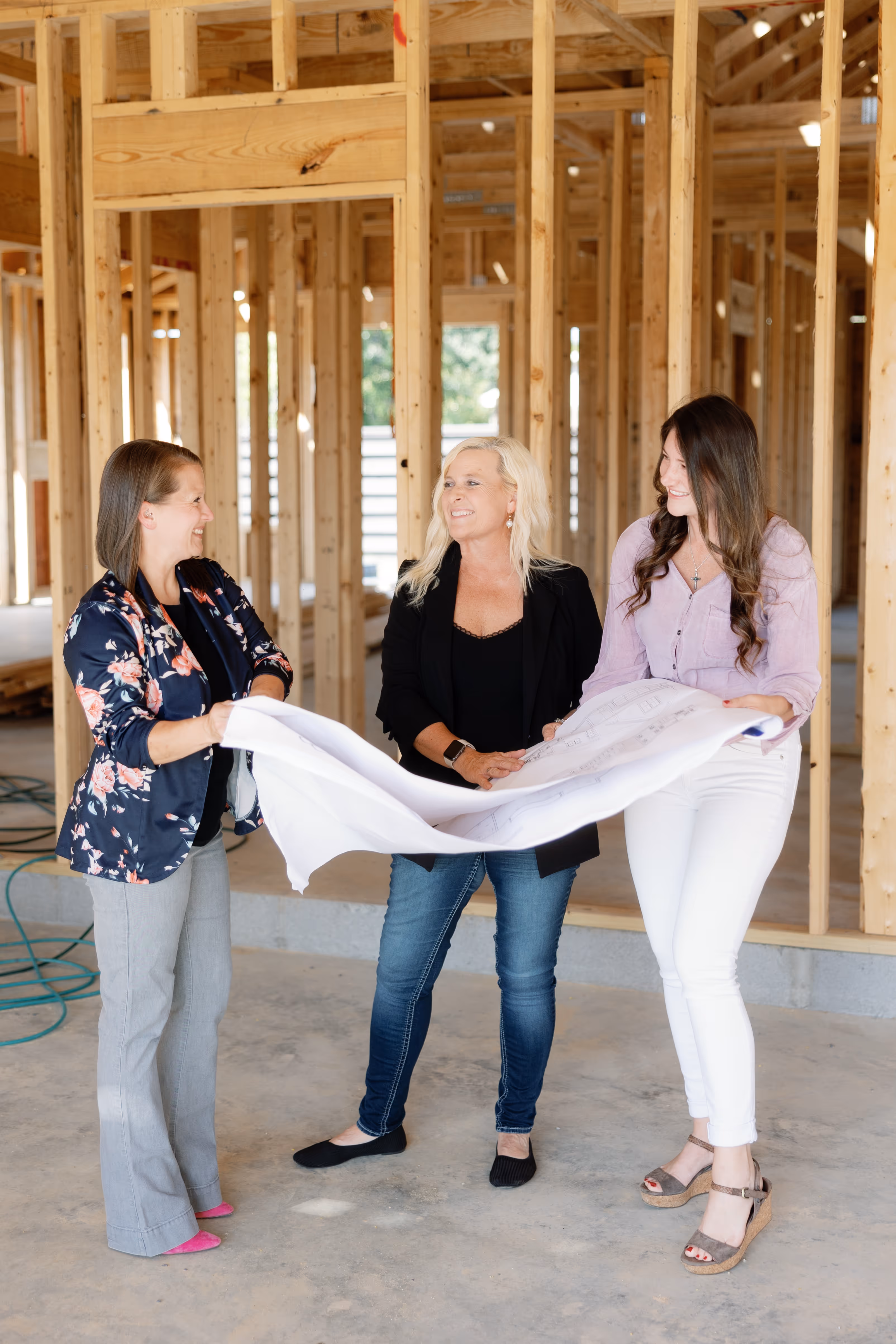 Three women standing inside a wooden house frame, holding and discussing a large blueprint.