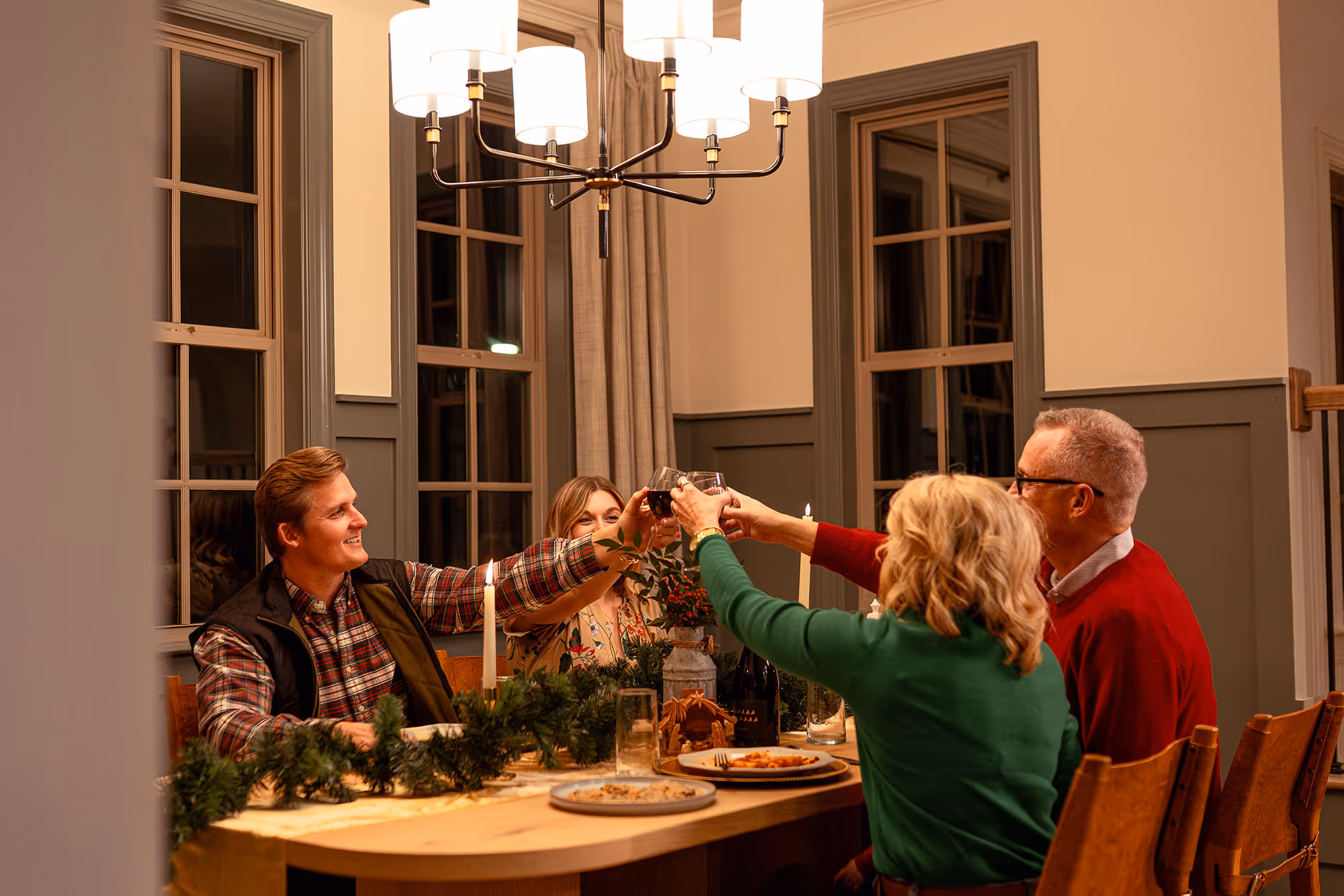 Four adults sitting around a dining table with holiday decorations, clinking wine glasses in a toast.