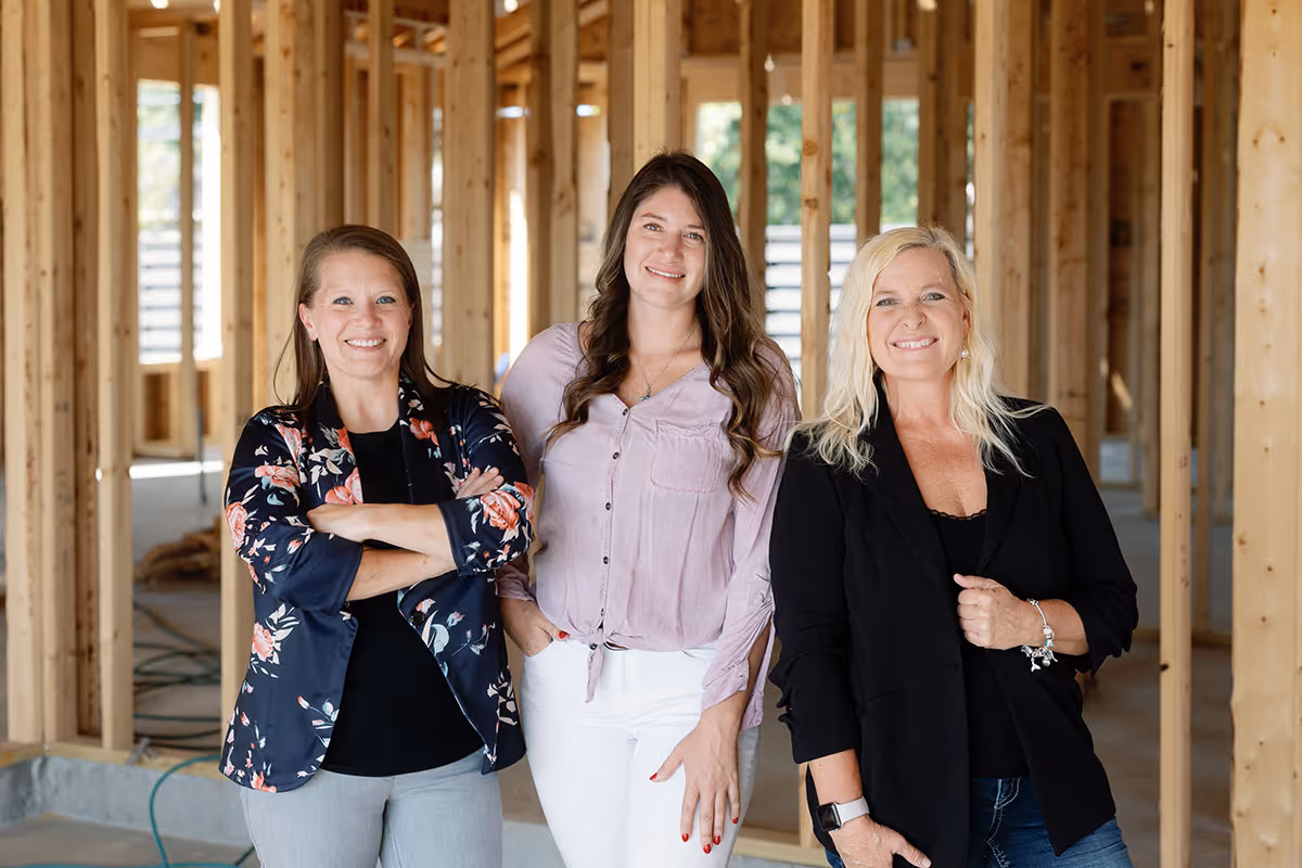 Three women standing and smiling inside a wooden framed house under construction.