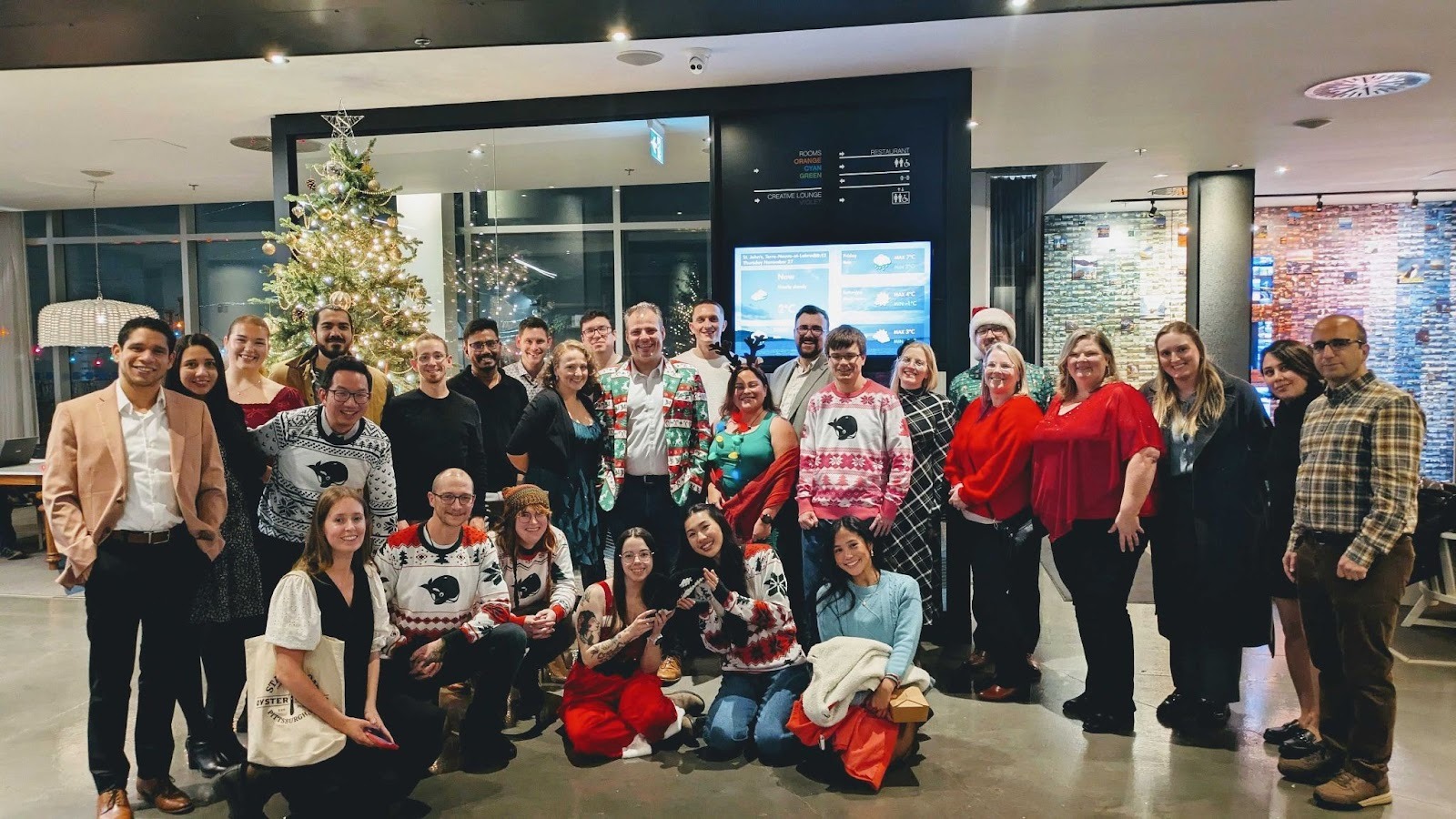 Group photo of HeyOrca employees gathering in a festive setting, with a decorated Christmas tree in the background. Attendees wear holiday-themed attire, including sweaters and festive colors. The atmosphere appears cheerful and celebratory.
