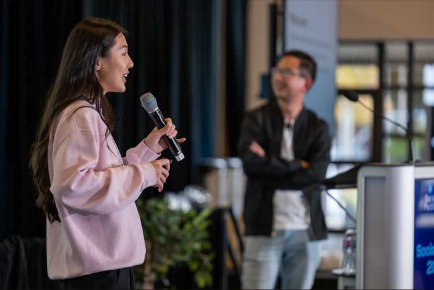 A woman in a pink sweater speaks into a microphone during a presentation, while a man in a black jacket stands in the background, listening attentively.