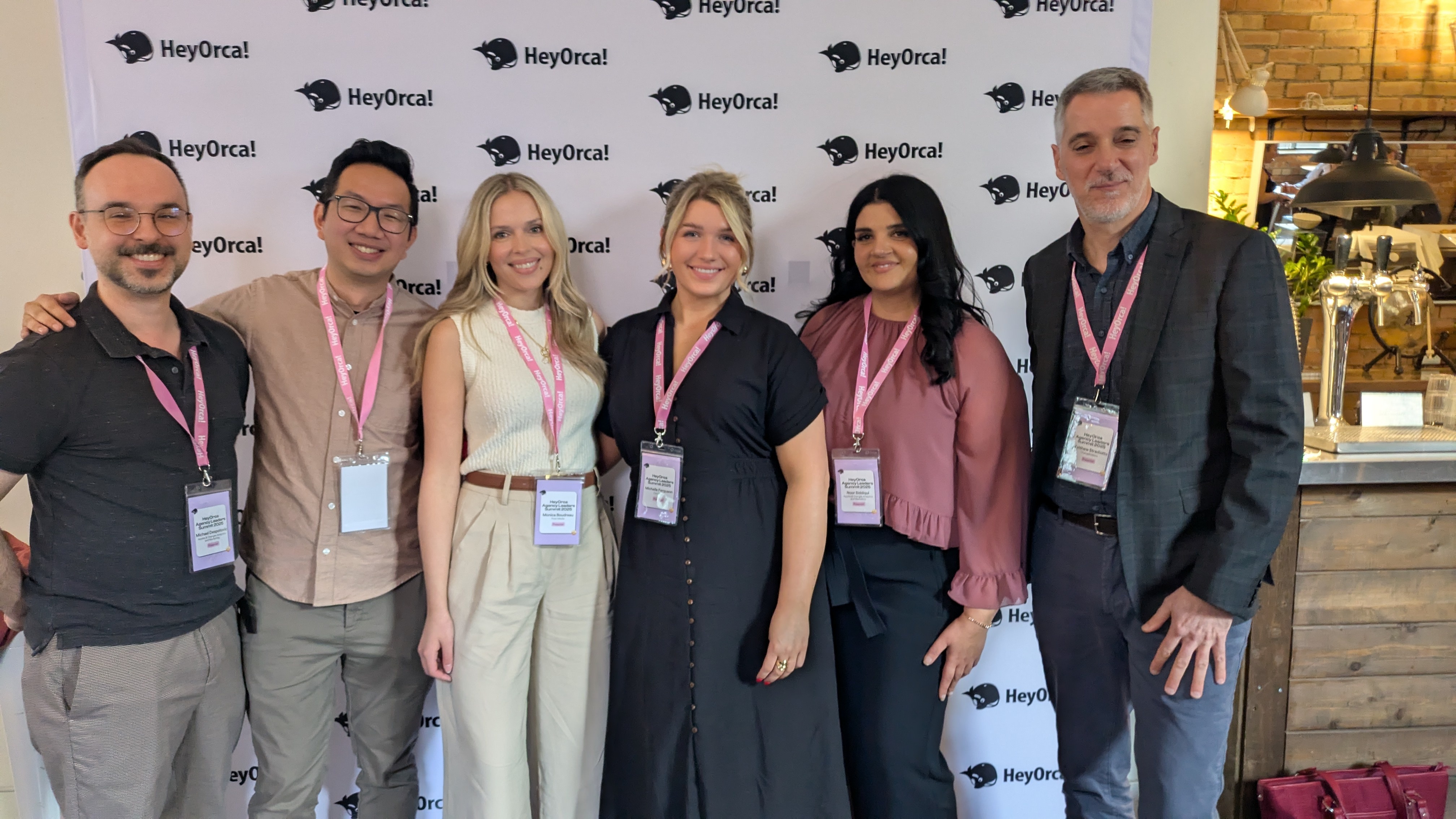 A group of six people stands smiling in front of a branded backdrop featuring the logo "HeyOrca." They are dressed in a mix of professional attire.