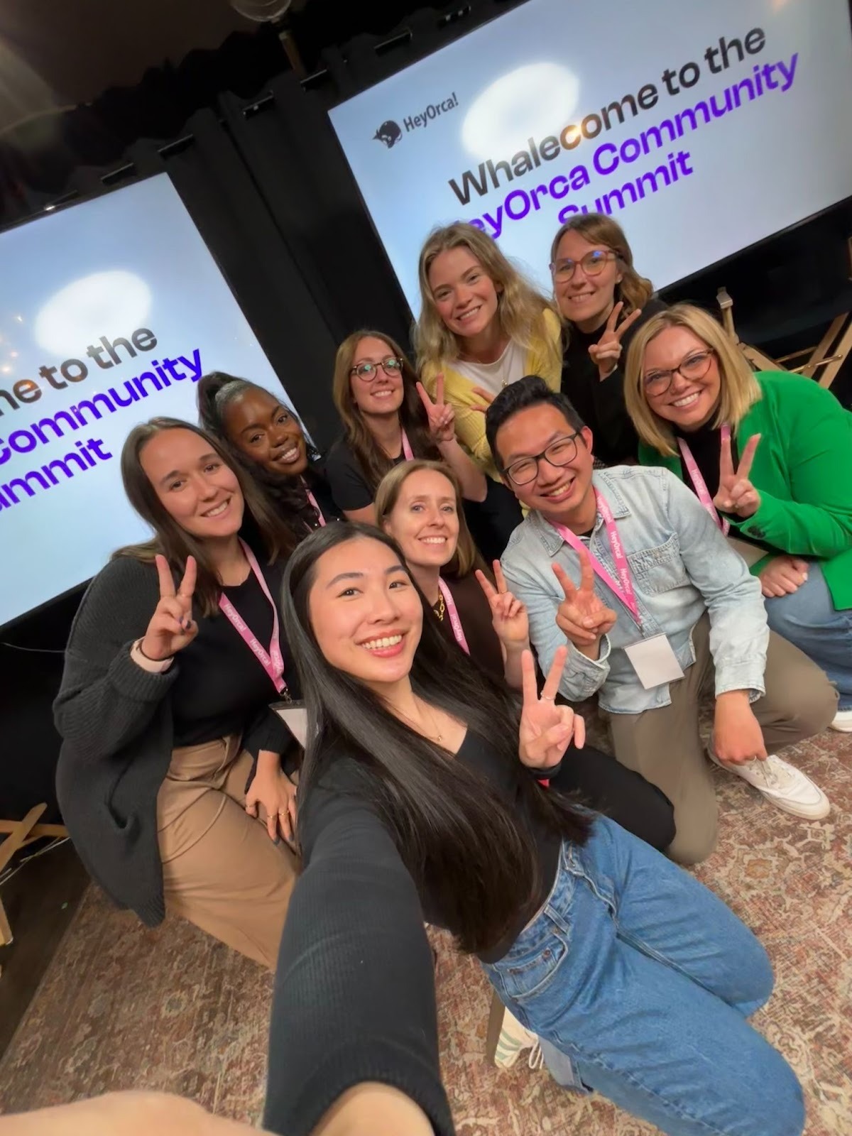 Group of nine individuals posing for a selfie at the HeyOrca Community Summit, smiling and making peace signs. Behind them are two large screens displaying the event's welcome message. The setting is an indoor venue with a warm atmosphere.