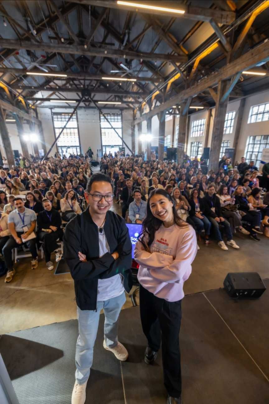 A diverse audience fills a large industrial space, seated and facing a stage. In the foreground, two speakers pose confidently: a man in a black jacket and a woman in a light pink sweatshirt. They smile, showcasing enthusiasm for the event.