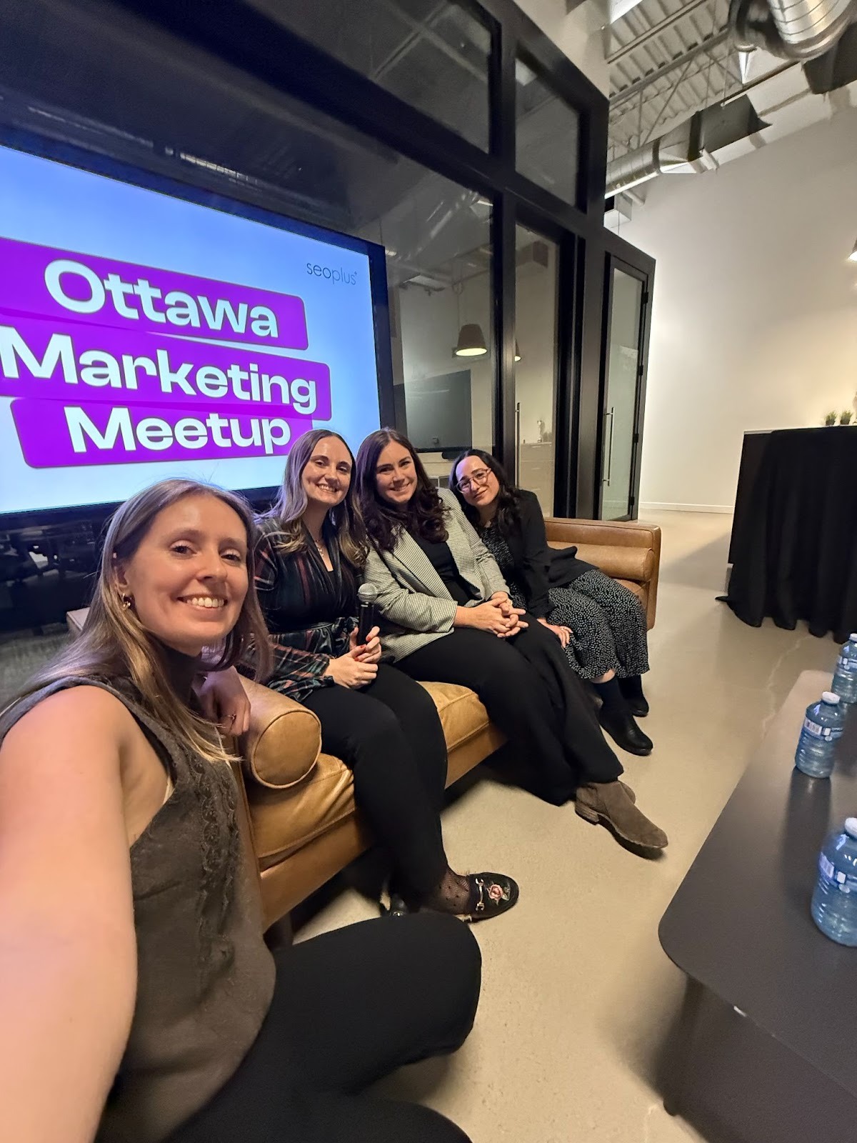 Four women sit on a couch at an Ottawa Marketing Meetup event, smiling at the camera. A large screen behind them displays the event name. The setting is a modern indoor space with a minimalistic design.