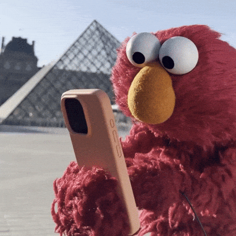 A red, fluffy puppet character is holding a smartphone while standing in front of the Louvre Pyramid. The background features the iconic glass structure of the museum against a clear sky.
