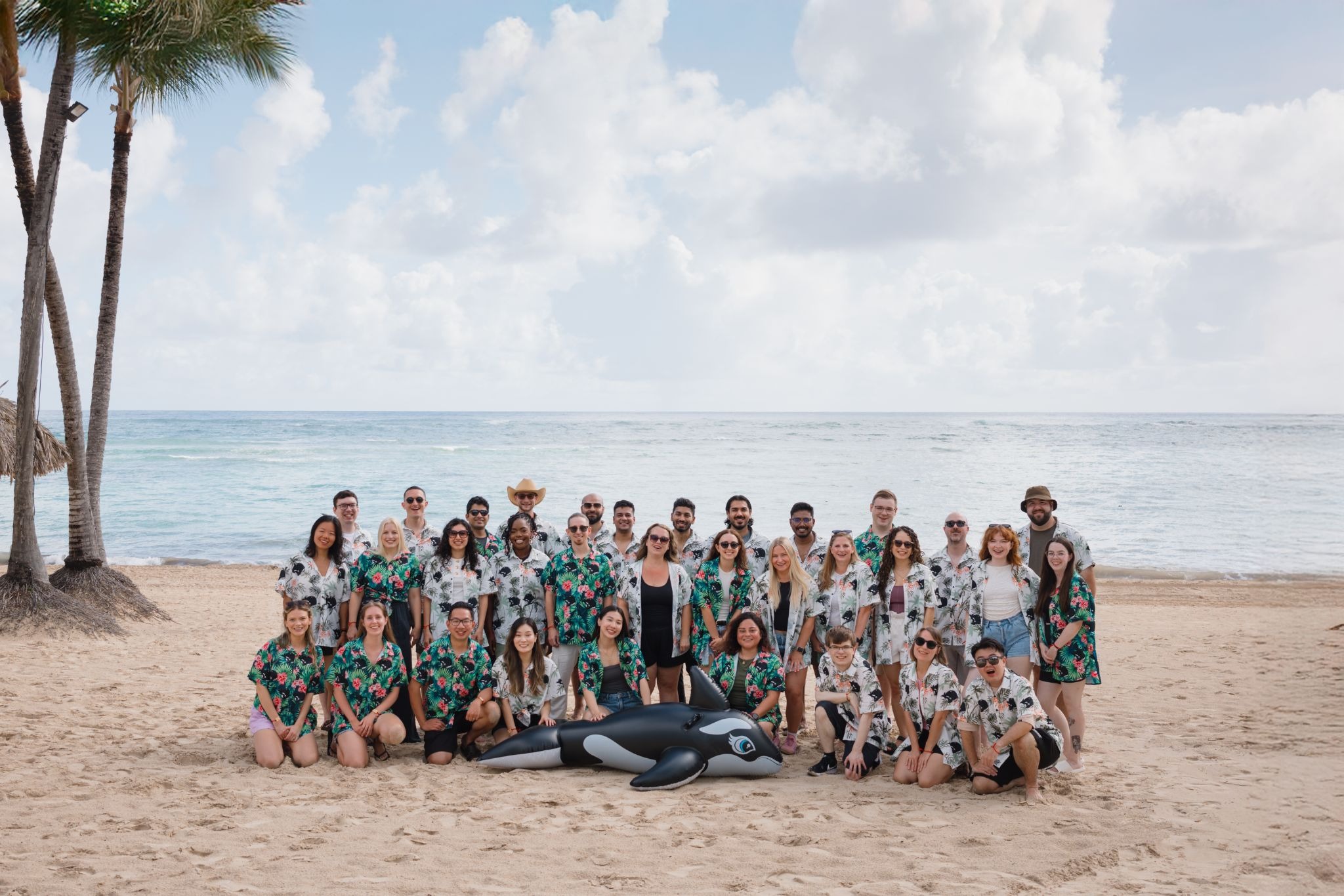 A large group of people stands on a sandy beach, wearing matching floral shirts. They are arranged in two rows, with a life-sized orca figure in front. Palm trees can be seen to the side, and the ocean stretches out behind them under a partly cloudy sky.