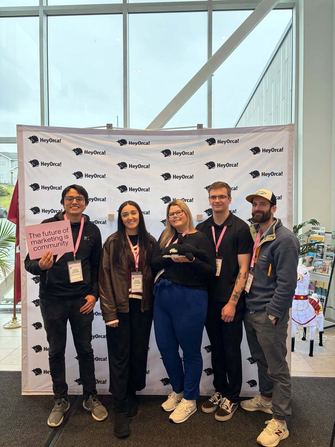  A group of five individuals stands together in front of a branded backdrop at an event. Three men and two women are present, all smiling. One man holds a sign that reads, "The future of marketing is community." The setting is bright with large windows in the background.