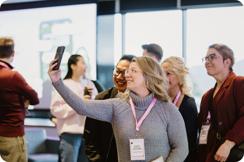 A group of people gathers in a modern setting, with one woman taking a selfie. She has long, blonde hair and wears a gray sweater. Others smile and pose behind her, showcasing a mix of casual and professional attire.