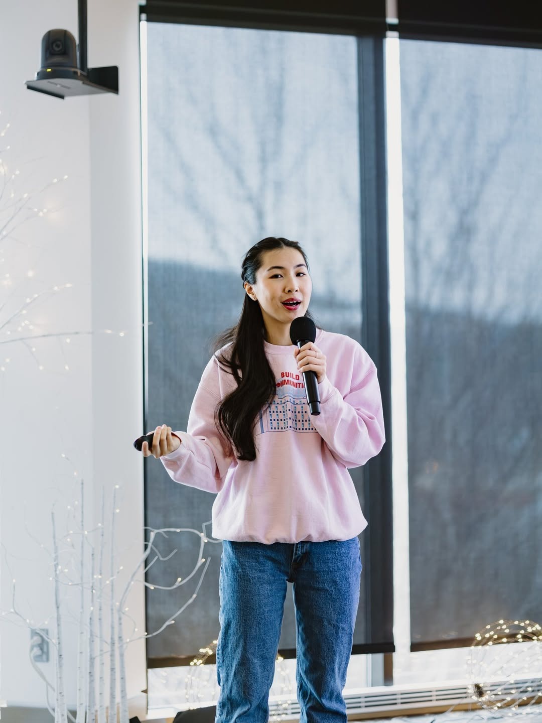  A young woman stands on a stage holding a microphone, wearing a pink sweatshirt and blue jeans. She appears to be speaking to an audience in a well-lit room with large windows. Soft lighting and decorative elements are visible in the background.