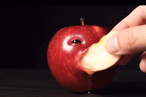 A hand holds a slice of apple, positioned to take a bite from a red apple that has an exaggerated facial expression and an eye. The background is dark, emphasizing the vibrant colour of the apple. Juice drips from the fruit as the slice approaches its mouth-like opening.