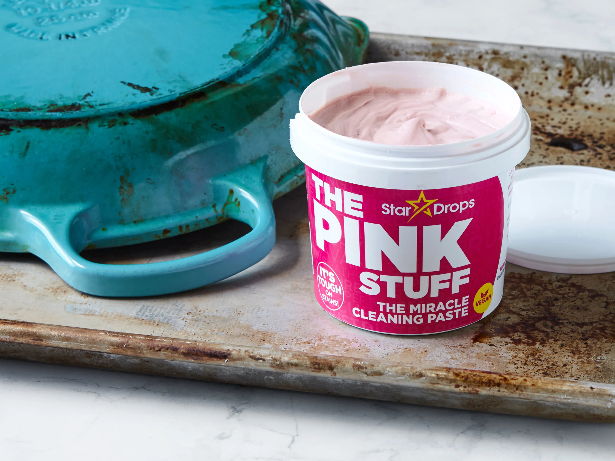 A tub of The Pink Stuff cleaning paste is open on a stained baking tray, next to a blue enamelled cast iron pot, showing its use for tough cleaning tasks.