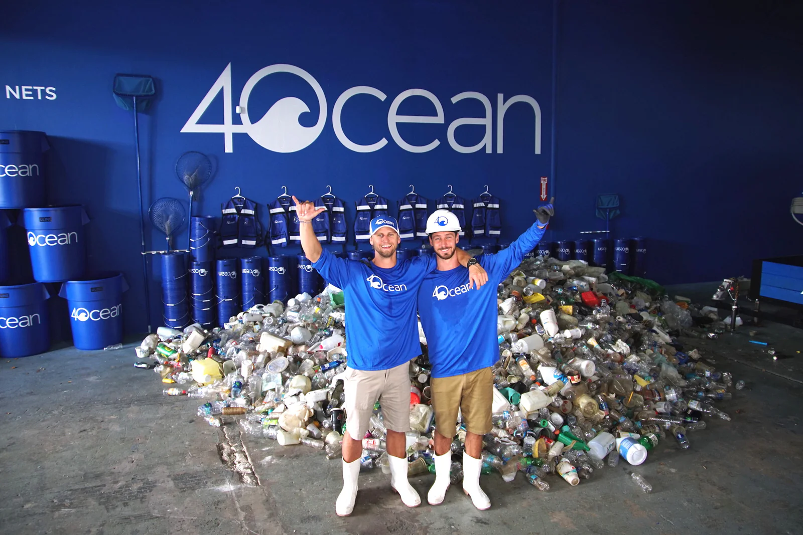 Two people wearing blue 4Ocean shirts stand in front of a large pile of collected plastic waste inside a 4Ocean facility, celebrating their cleanup efforts.