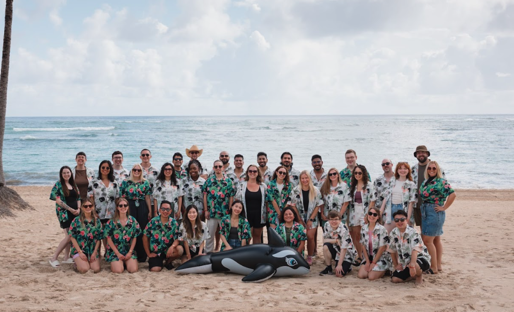 A large group of people stand together on a sandy beach, with a scenic ocean view in the background. Many wear matching floral shirts, and a large inflatable orca lies in front of the group.