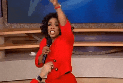 An energetic speaker in a bright red suit raises their arm on a studio stage, holding a microphone. There's a blue screen in the background.