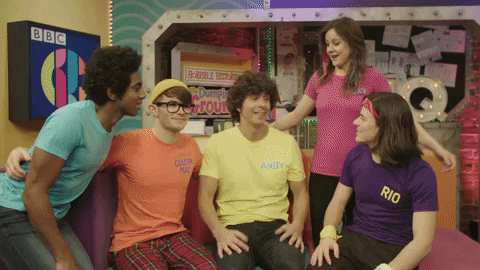 A group of five people sitting on a purple couch, each wearing brightly colored shirts with their names printed on them. They appear to be in a lively discussion, smiling and interacting with each other.