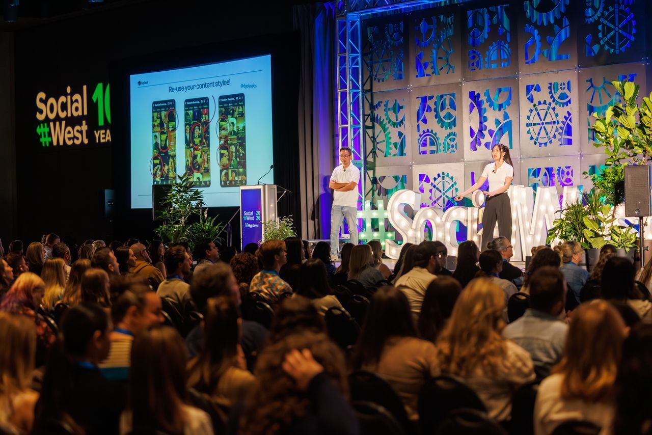 Two speakers address a large audience from a stage at a conference. Behind the speaker, a screen displays a colourful presentation. The event is titled 'SocialWest' with decorative stage elements and plants.
