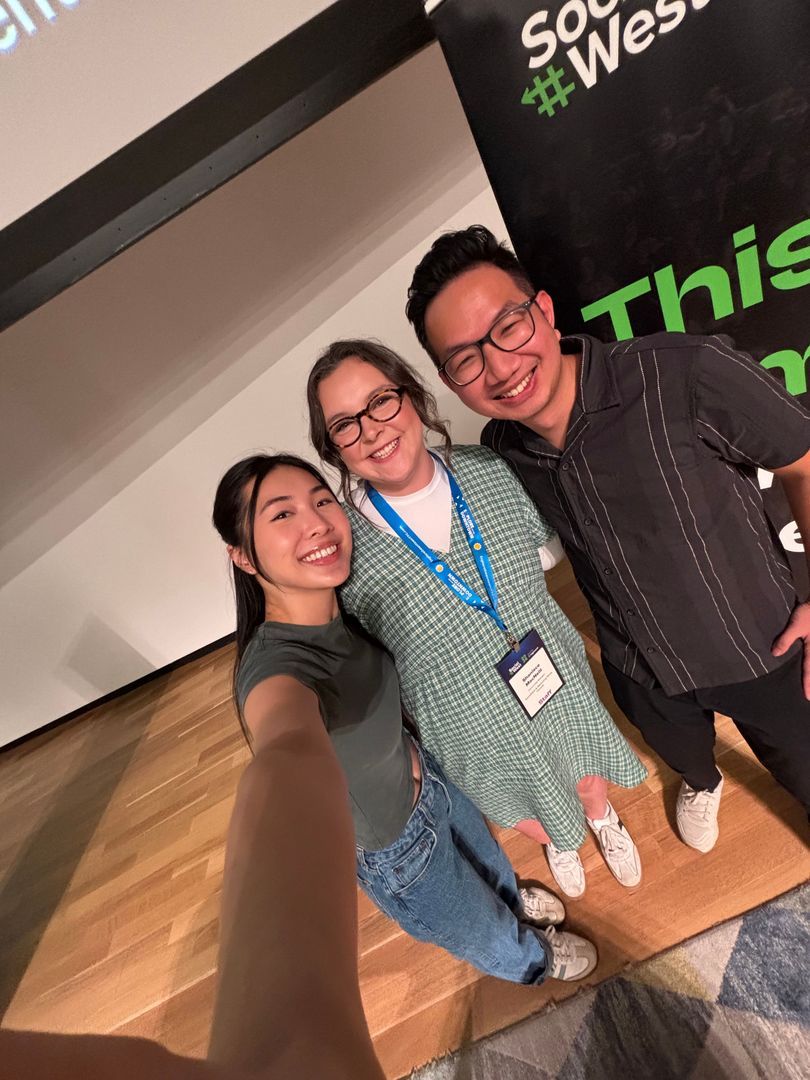 Three people posing for a selfie in front of a stage backdrop at a conference. They are smiling and standing close together.