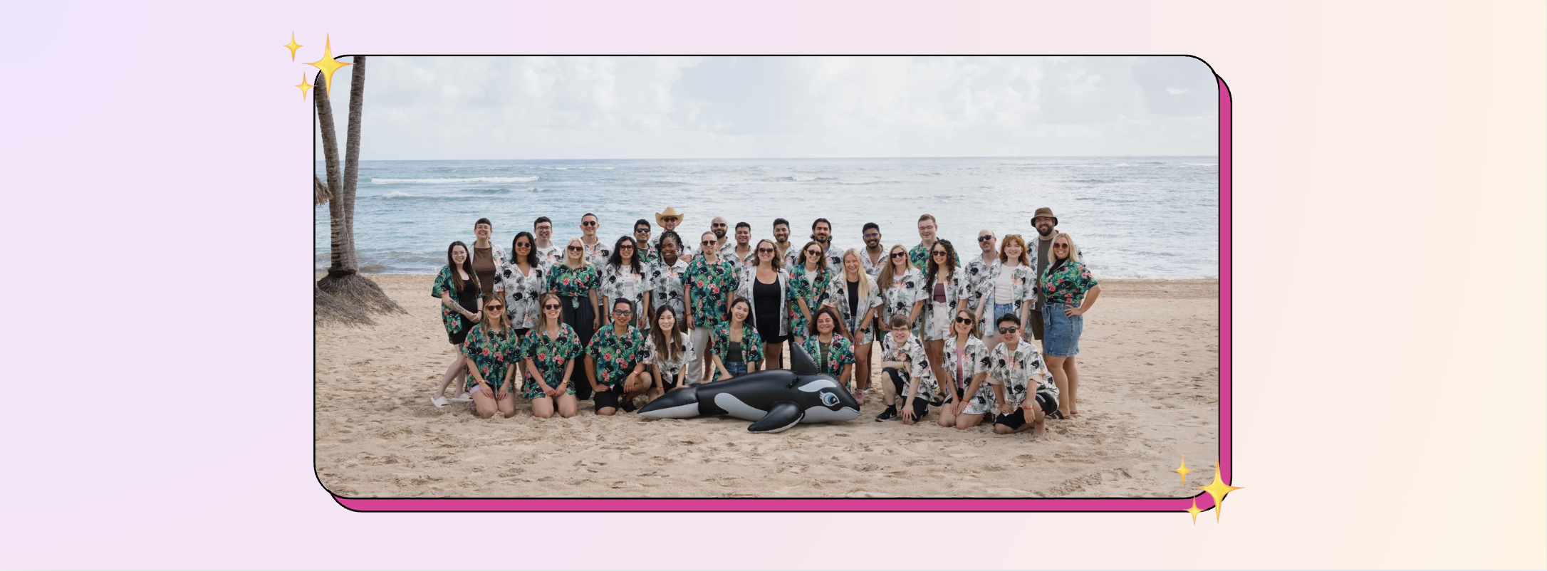 A large group of people in matching tropical shirts pose on a sandy beach with a scenic ocean backdrop. They are gathered around inflatable orcas, smiling for the camera.