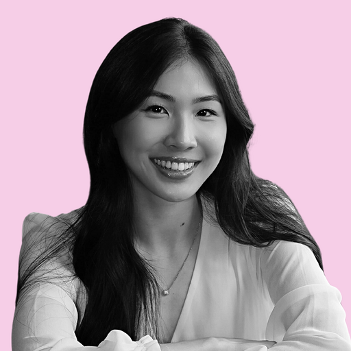 A woman with long dark hair sits with her arms crossed on a pink background, wearing a light blouse and smiling gently at the camera.