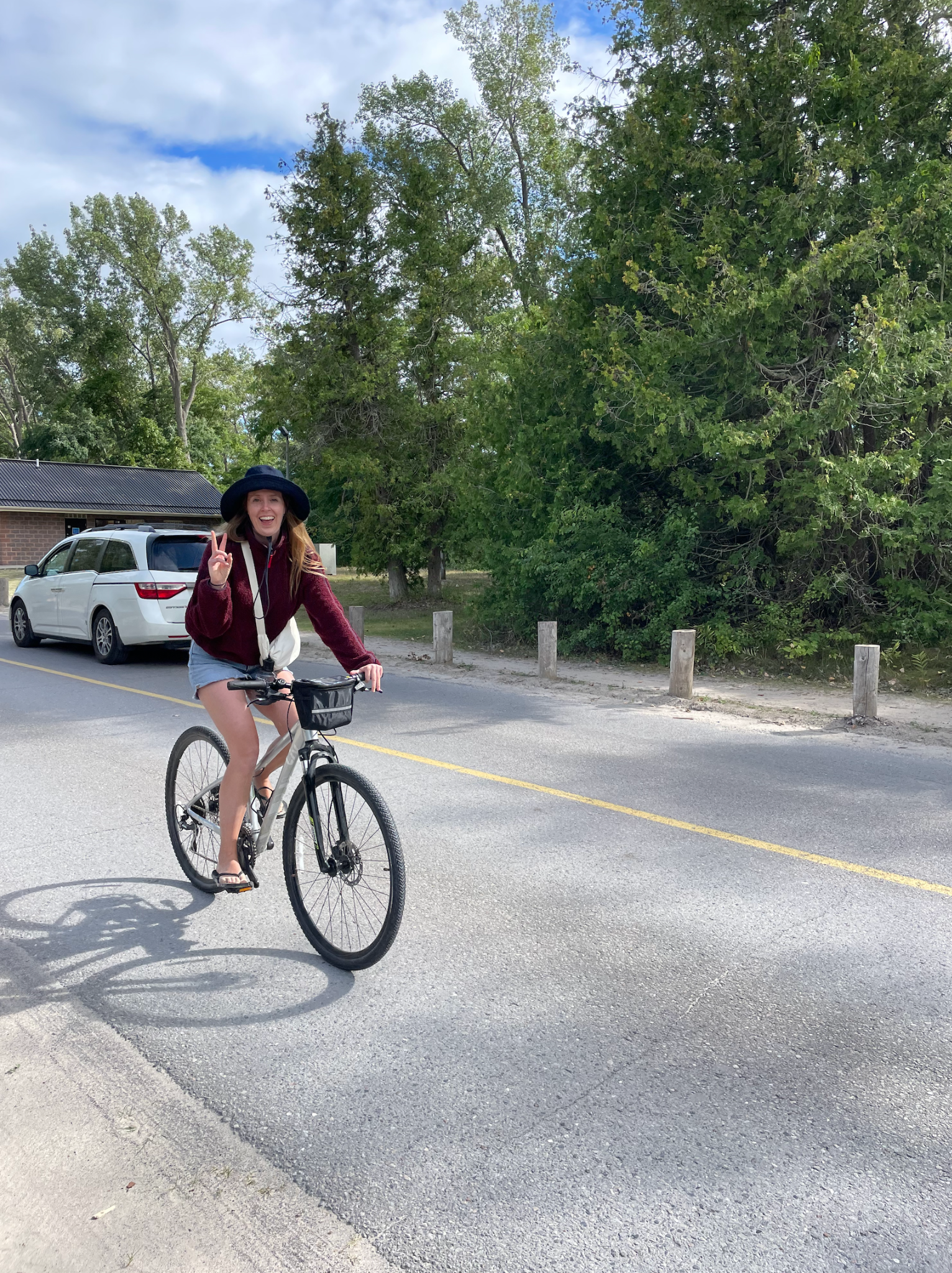 A woman wearing a large hat and casual clothing rides a bike on a suburban road. Trees and a parked car are visible in the background.
