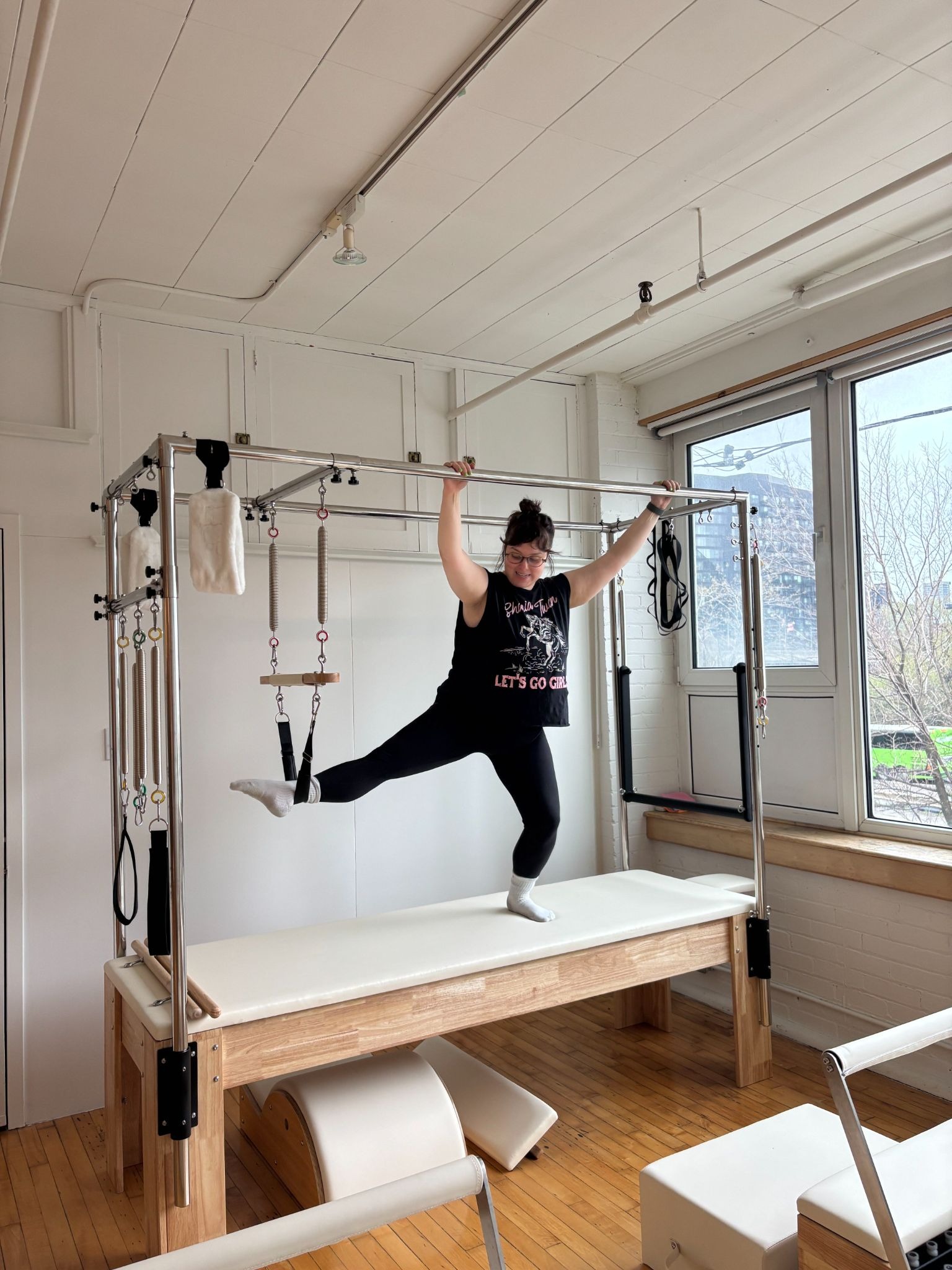 A person practices on a Pilates Cadillac reformer in a bright studio, balancing on one leg with arms extended.