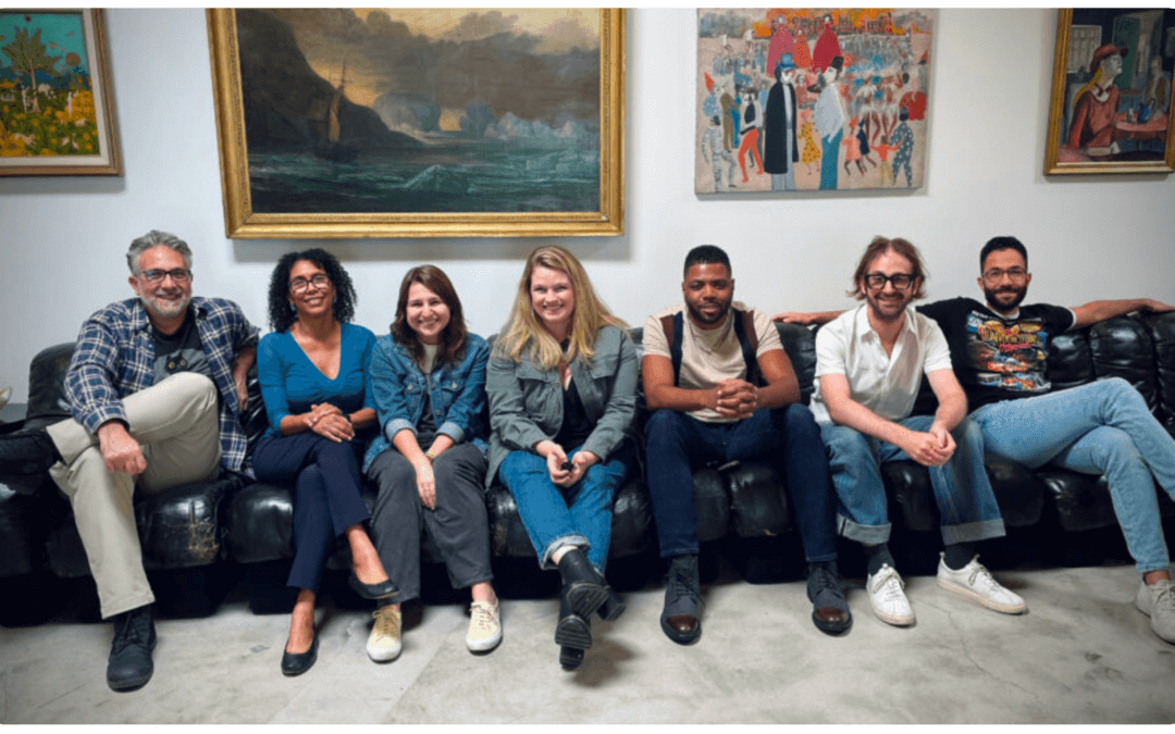 A group of seven people sitting on a leather couch in an art gallery with several paintings on the wall behind them. They are casually dressed and smiling.