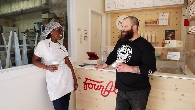 Two people are in a modern ice cream shop setting. The woman on the left wears a work uniform, indicating she may be a staff member. The man on the right is dressed casually. They are having a relaxed discussion.
