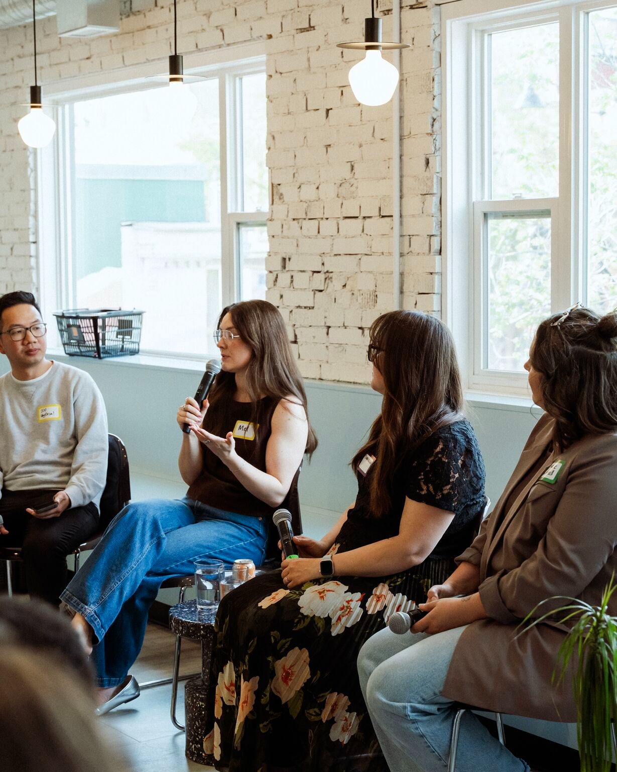 A panel of four people sit in a well-lit room with brick walls and large windows. Three women holding microphones are engaged in conversation, while a man in a sweater listens attentively.