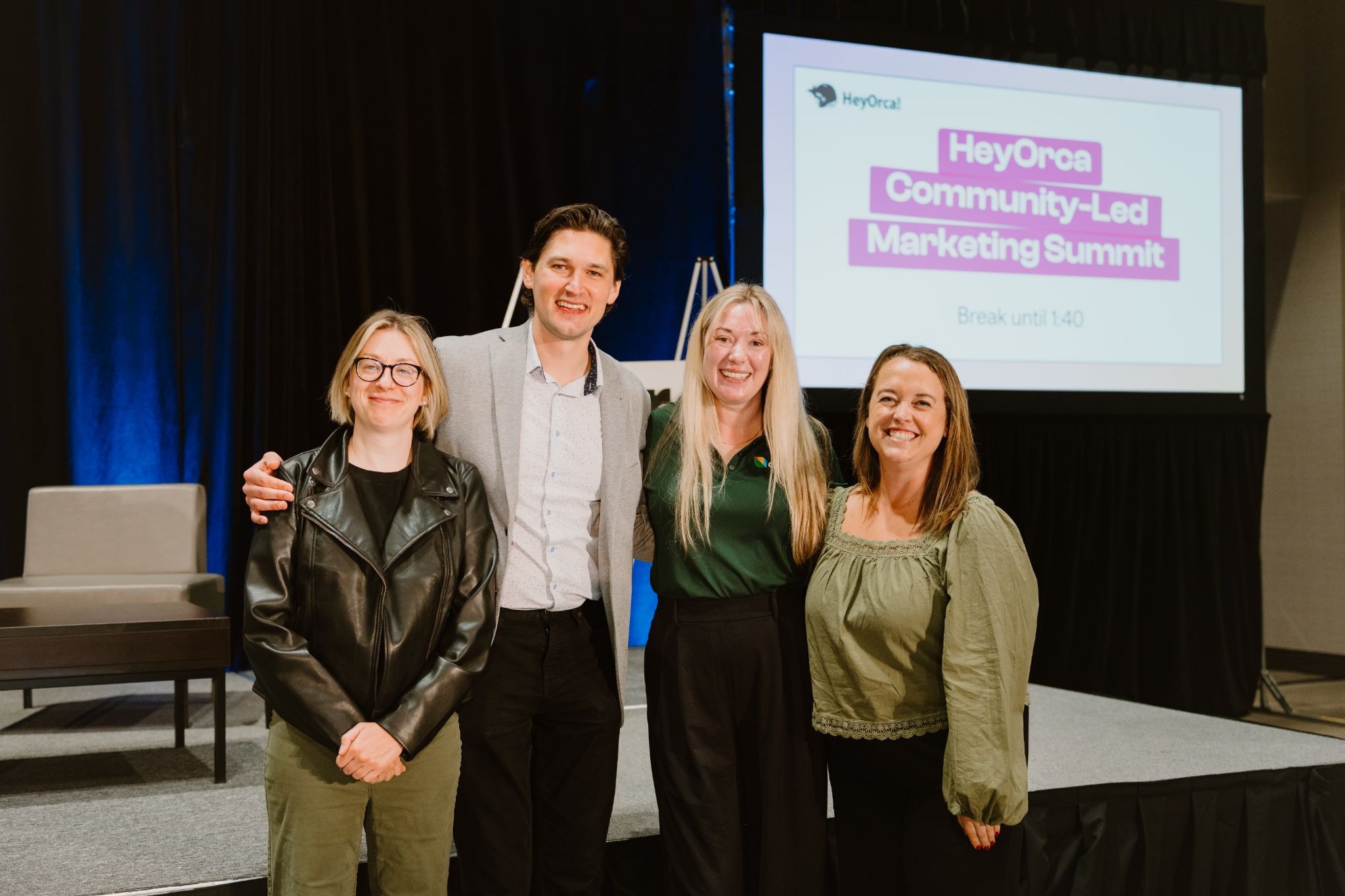 Four people standing together and smiling at the HeyOrca Community-Led Marketing Summit. A sign with the event name is visible in the background.