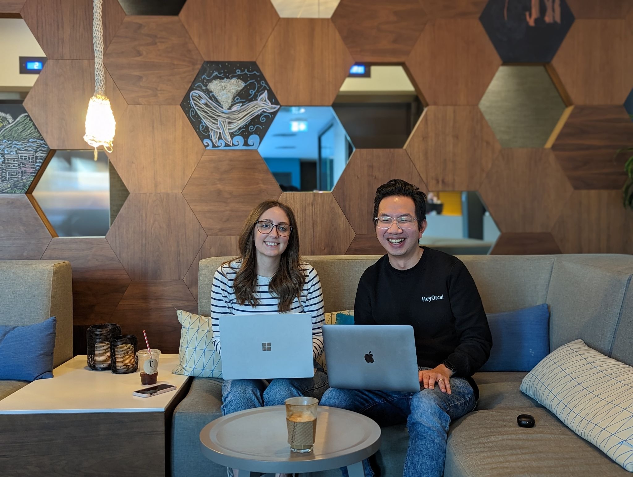Two people sitting on a sofa with laptops in a modern office space featuring hexagonal wall tiles and soft lighting. They appear to be working collaboratively.