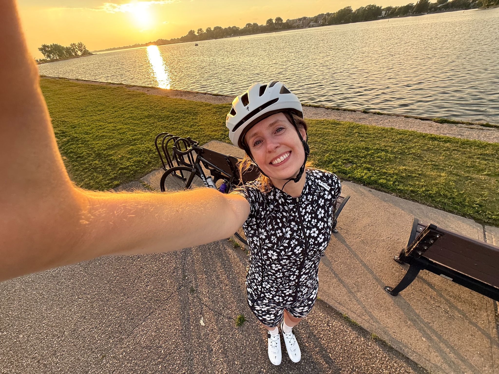 A person wearing a floral cycling outfit and a bicycle helmet takes a selfie by a lakeside path during sunset. Bicycles are parked next to benches along the shoreline.