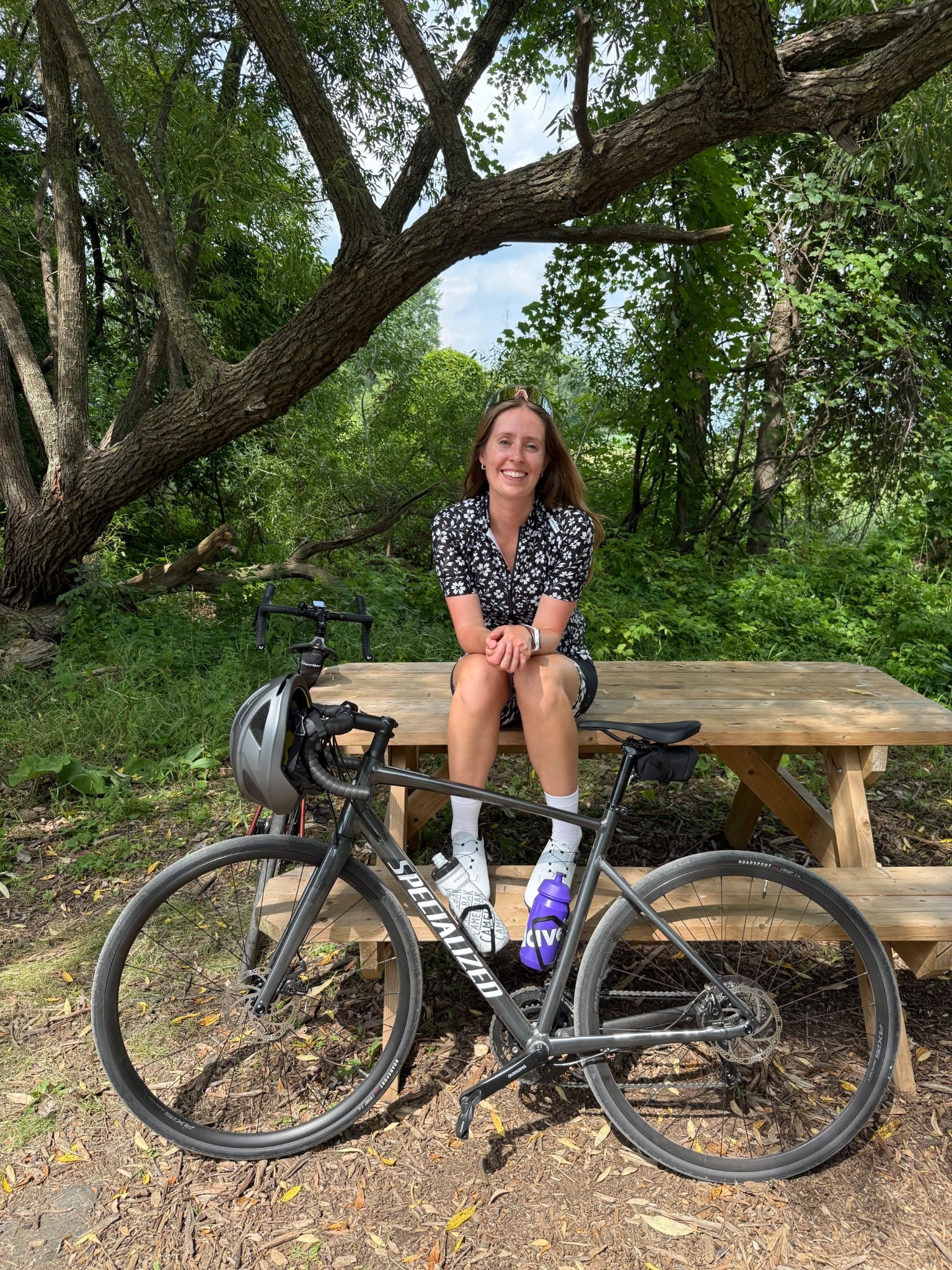 A person sitting on a picnic table in a park with a bicycle parked in front. The individual is smiling and wearing a casual outfit and cycling shoes.