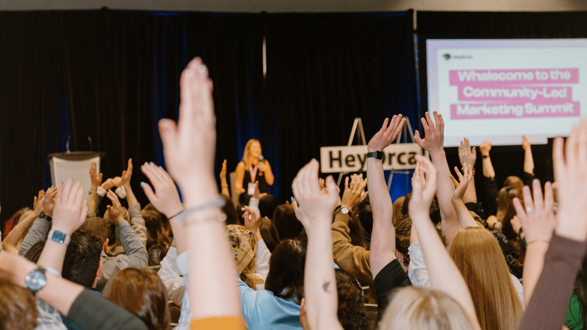 Audience members raise their hands during a presentation at a marketing summit. A screen displays a welcoming message.
