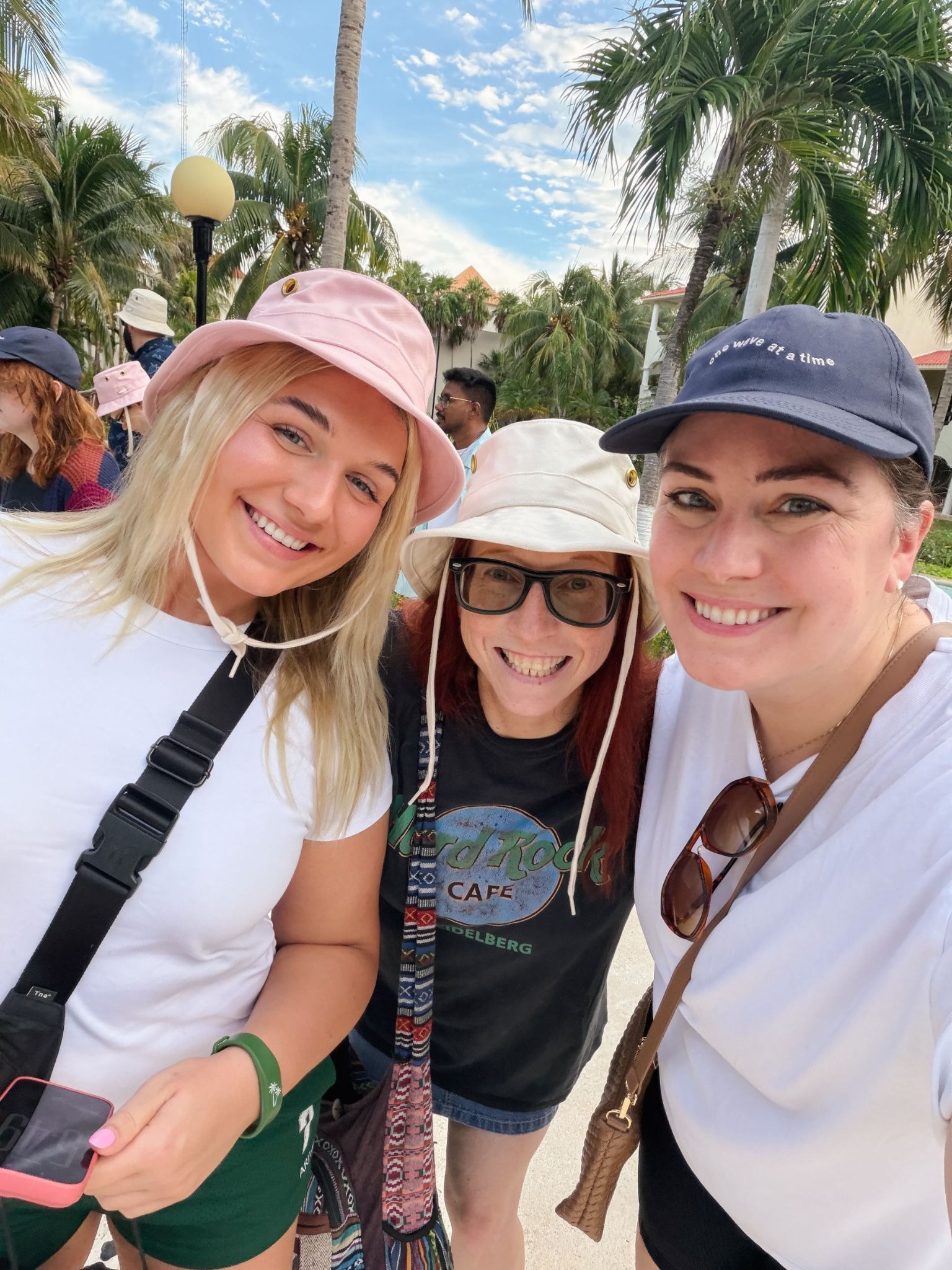 Three friends smiling and wearing hats, posing together outdoors with palm trees in the background, suggesting a tropical or vacation setting.