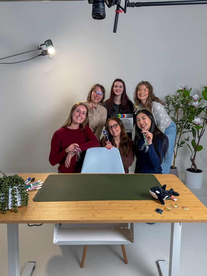 Six people standing behind a wooden desk with crafting materials, posing in a well-lit studio setting.