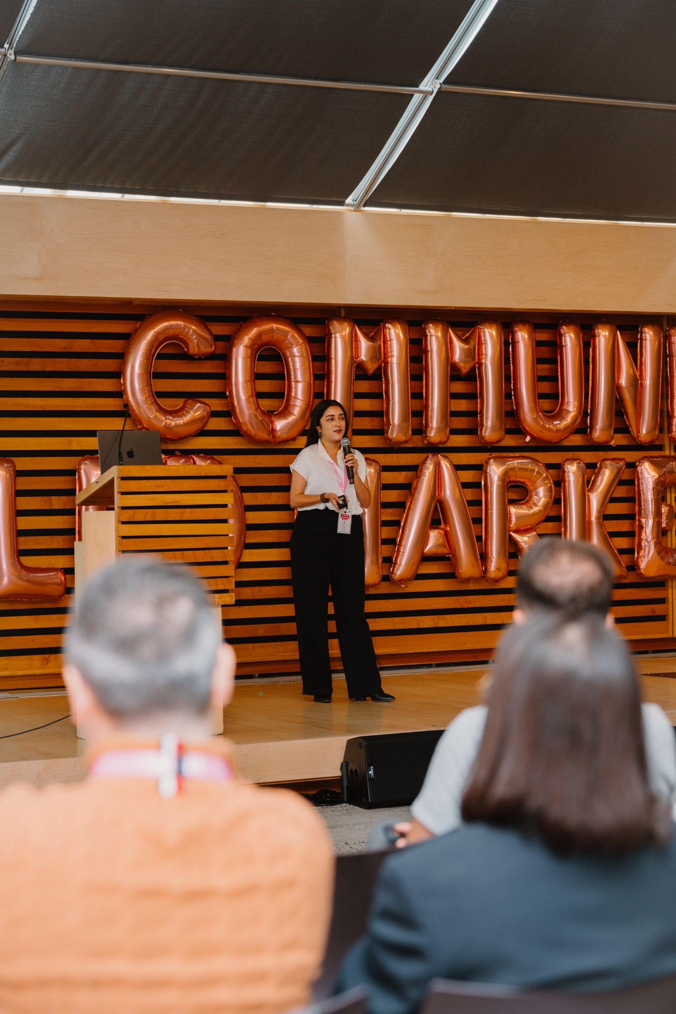 A speaker stands on stage addressing an audience. Large, metallic balloon letters spelling 'COMMUNITY' are in the background. The focus is on the speaker, engaging with the seated attendees.