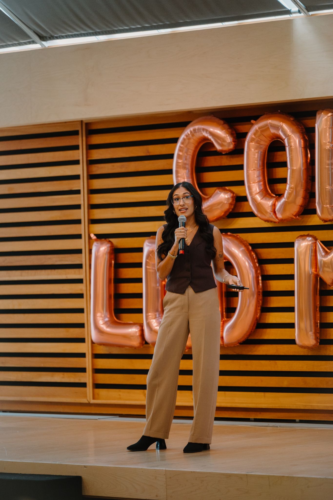 A person stands on a stage with a microphone, wearing a brown vest and tan pants. Behind them, large, metallic balloon letters are partially visible against a wooden slat wall.