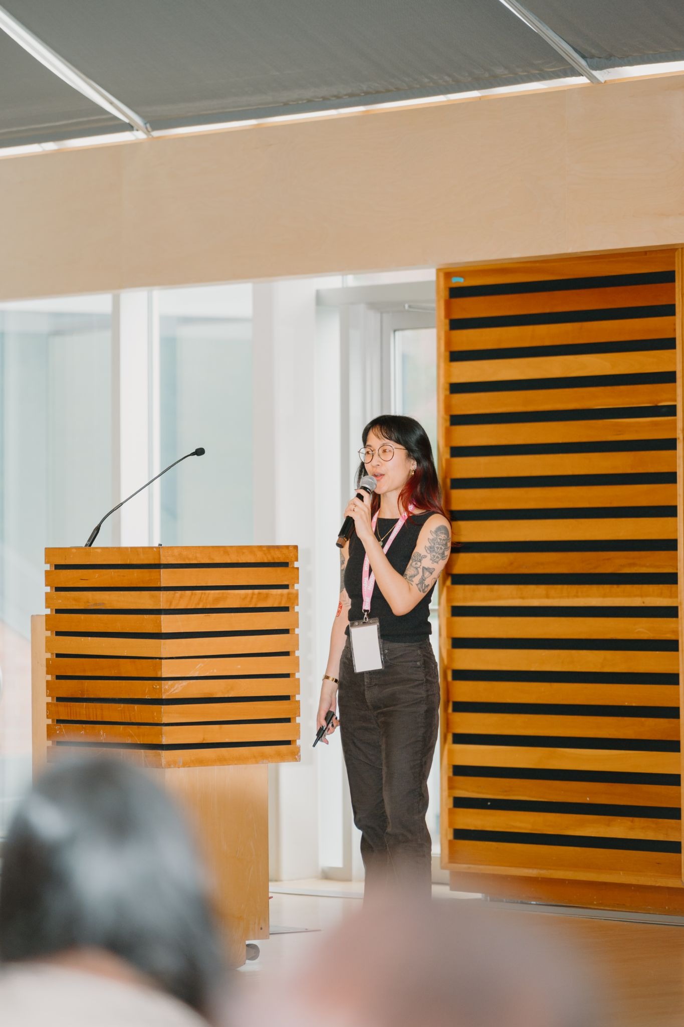A person giving a presentation on stage, standing beside a wooden podium. The room has large windows and modern decor.