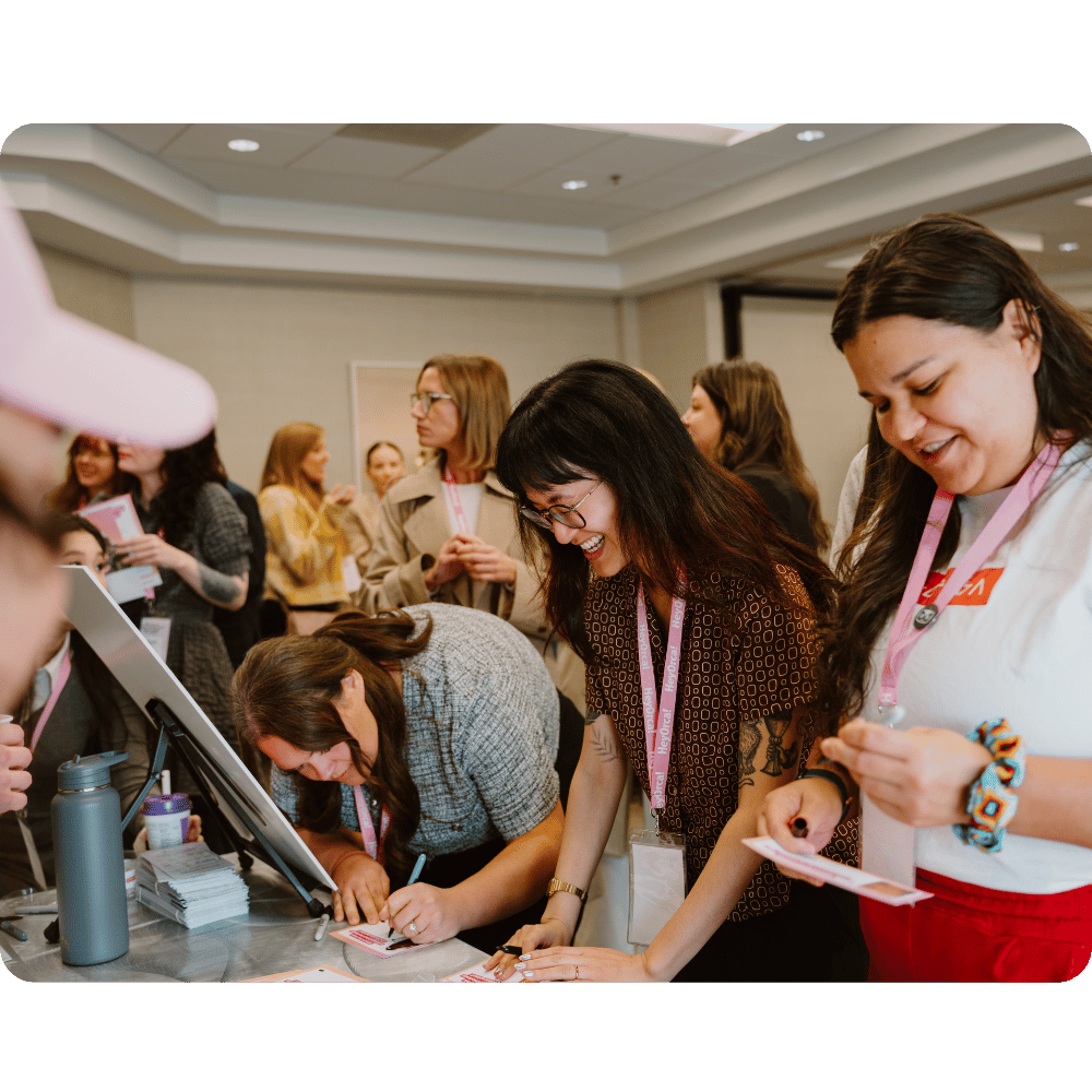 Women gathering at a conference registration area, filling out forms and receiving name tags. The atmosphere is lively, with attendees interacting and preparing for the event.