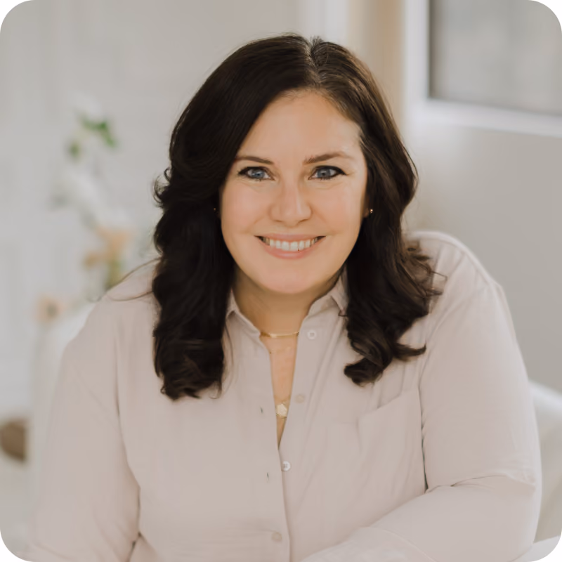 A woman with dark hair smiles for the camera. She wears a cream button up top.