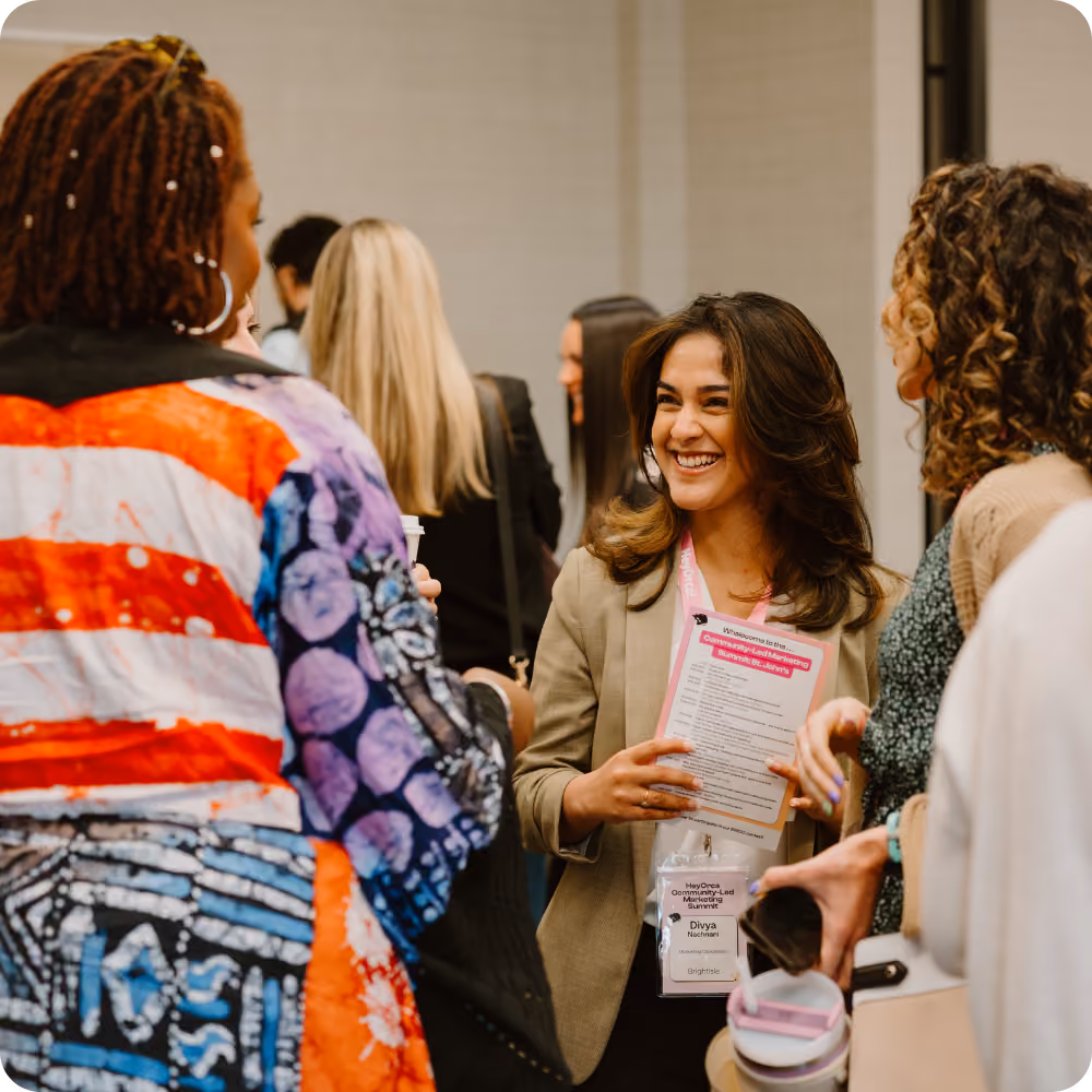 A diverse group of women engaged in conversation at a professional networking event. One woman holds a schedule, and all appear engaged and smiling.