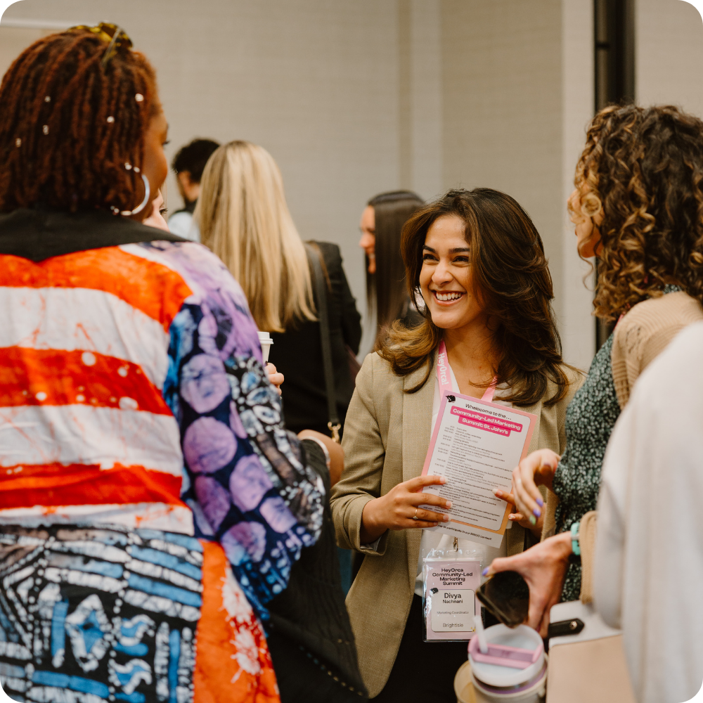 A diverse group of women engaged in conversation at a professional networking event. One woman holds a schedule, and all appear engaged and smiling.