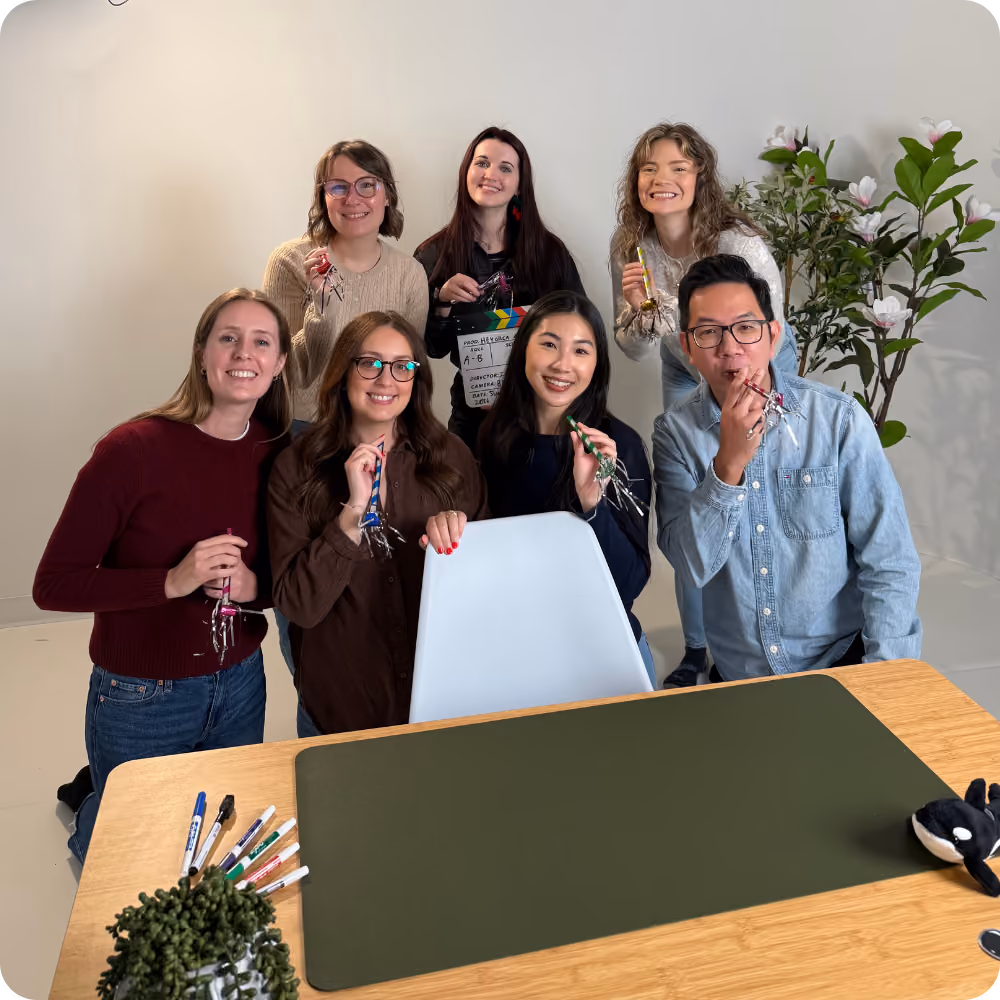 A group of seven people stand around a desk, smiling at the camera. They hold small noise makers, indicating a celebratory mood. The desk is equipped with office supplies, and there's a potted plant in view.