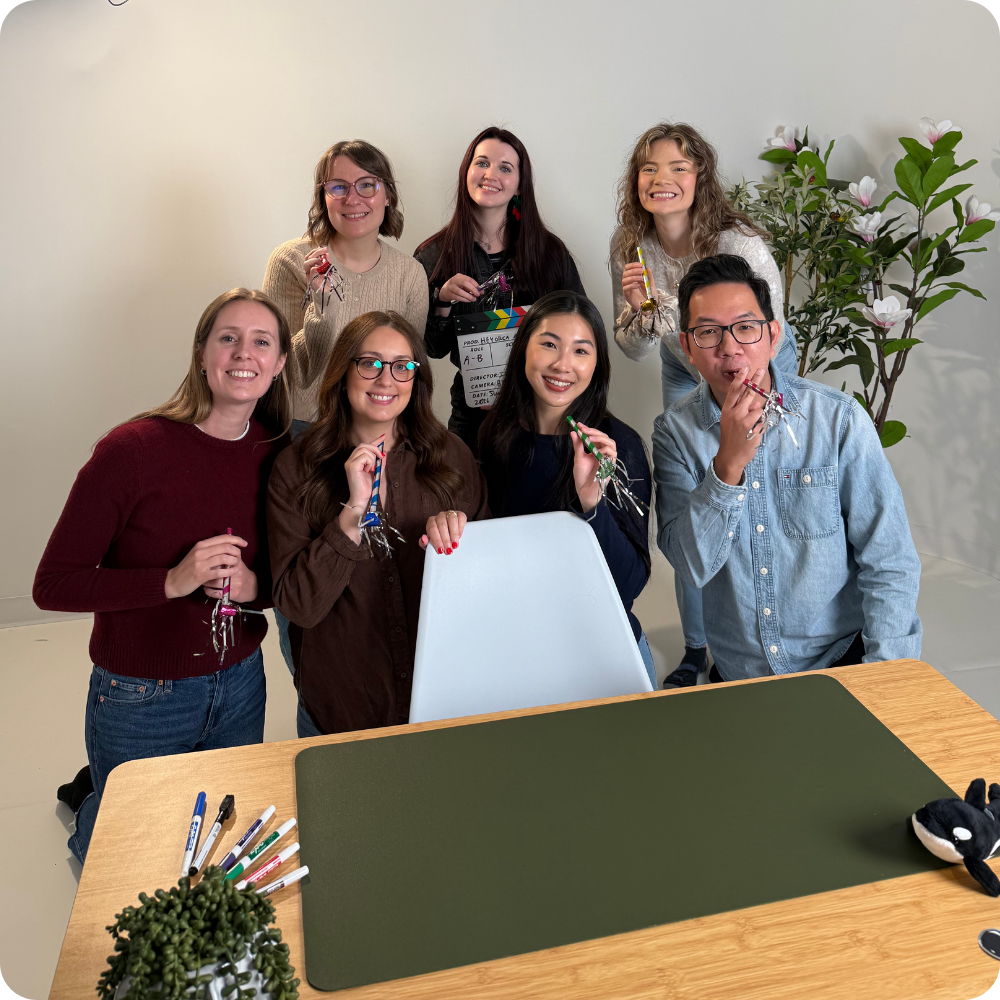 A group of seven people stand around a desk, smiling at the camera. They hold small noise makers, indicating a celebratory mood. The desk is equipped with office supplies, and there's a potted plant in view.