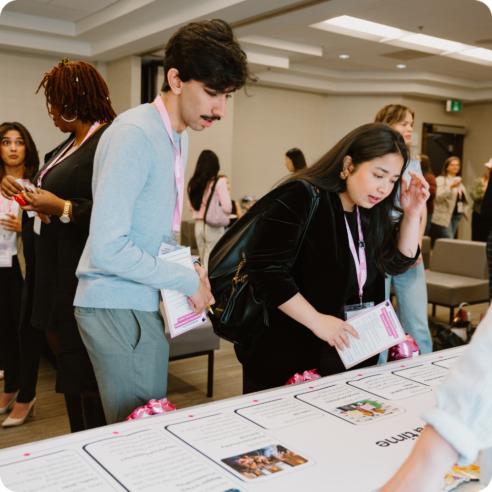 A group of attendees at a conference are gathered around a table, reviewing documents. The table displays various informational sheets and flyers. The participants are wearing lanyards, suggesting an organized event setting.